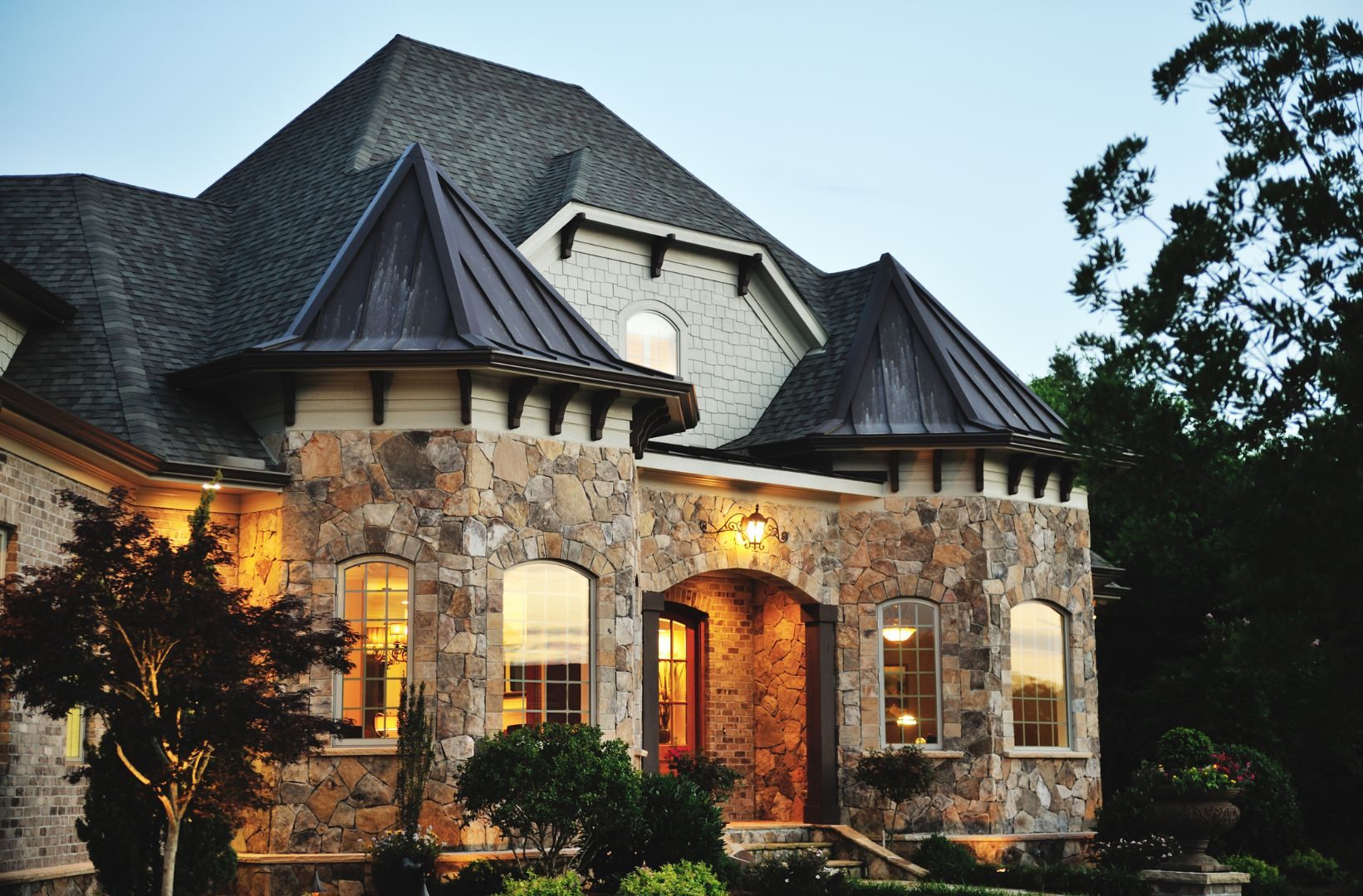 Large stone house with dark roof, illuminated windows, and a well-manicured garden, seen at dusk.