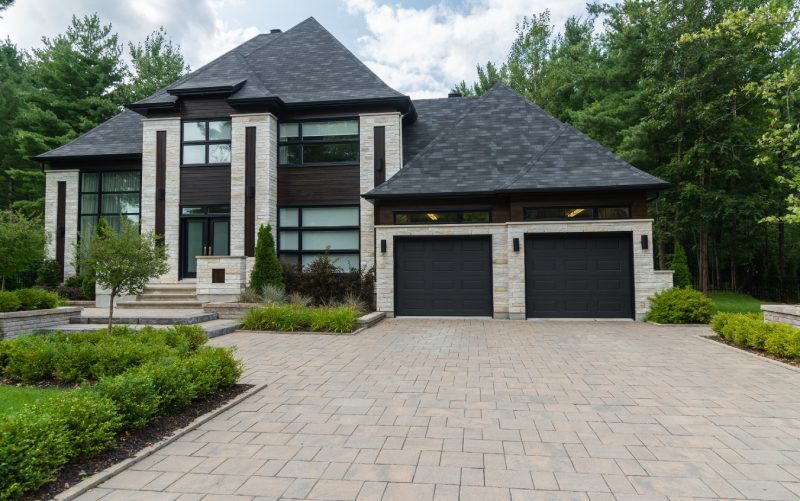 A modern two-story house with dark roofing, large windows, and a double garage. The front yard features a paved driveway and well-maintained landscaping, surrounded by trees.