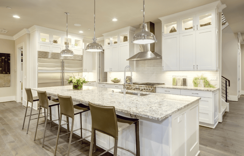 A modern kitchen features a large island with granite countertops, four barstools, stainless steel appliances, white cabinets, hanging light fixtures, and under-cabinet lighting.