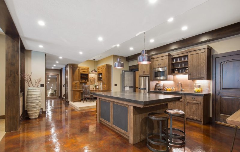 A spacious kitchen features wooden cabinets, a large island with two bar stools, pendant lighting, stainless steel appliances, and a polished brown floor. There is a dining area in the background.