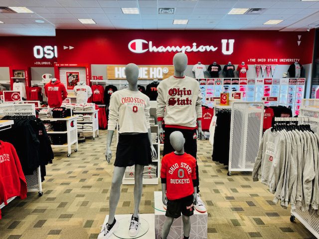 A store display featuring mannequins dressed in various Ohio State University apparel. The backdrop includes shelves filled with branded shirts, hoodies, and accessories.