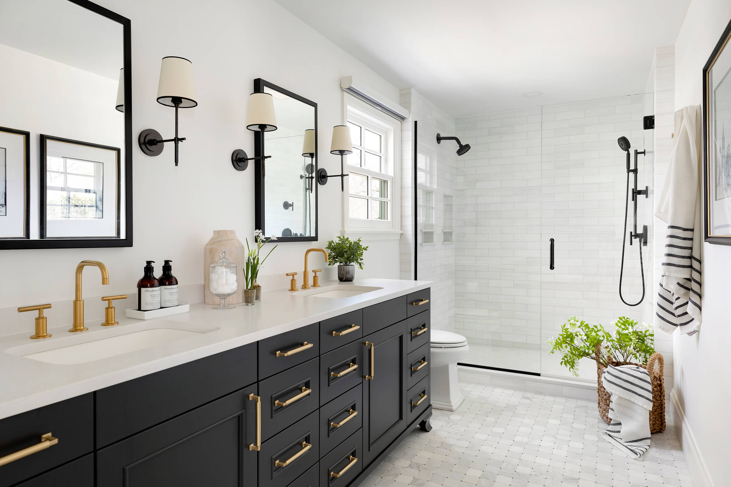 Modern bathroom with double vanity, gold fixtures, and black accents. Glass-enclosed shower in the background. White walls, large mirrors, and indoor plants enhance the space.