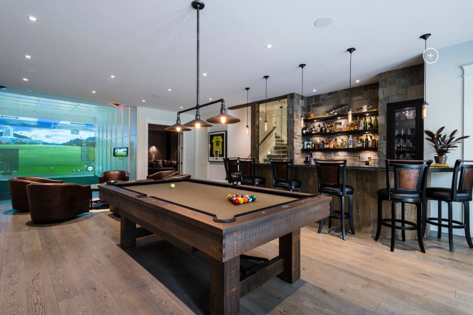 A game room with a pool table, bar, and seating area. A large screen displays a virtual golf course.