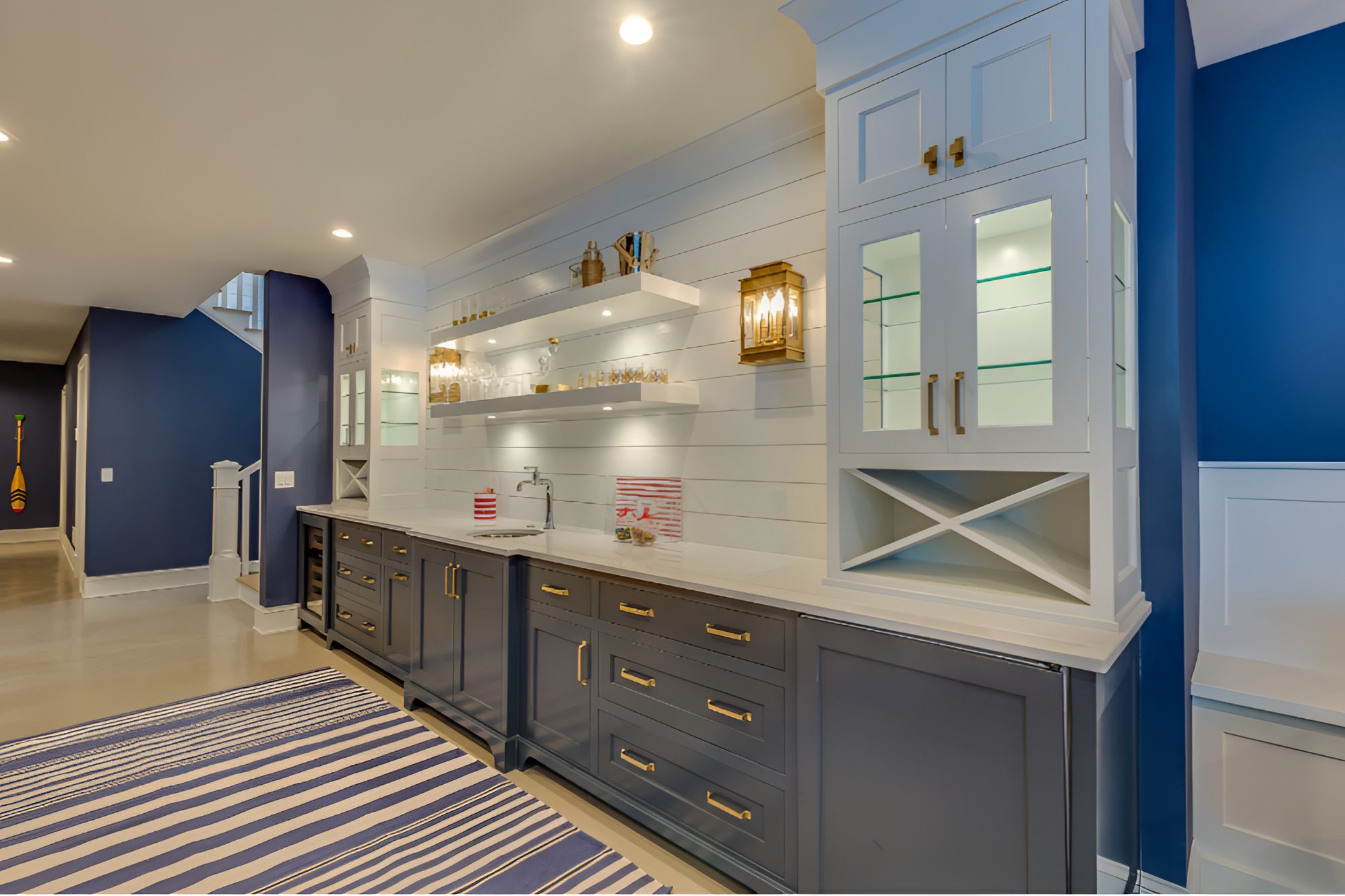 A modern home bar with navy blue cabinets, white countertops, gold accents, and open shelving. A striped rug lies on the light-colored floor.