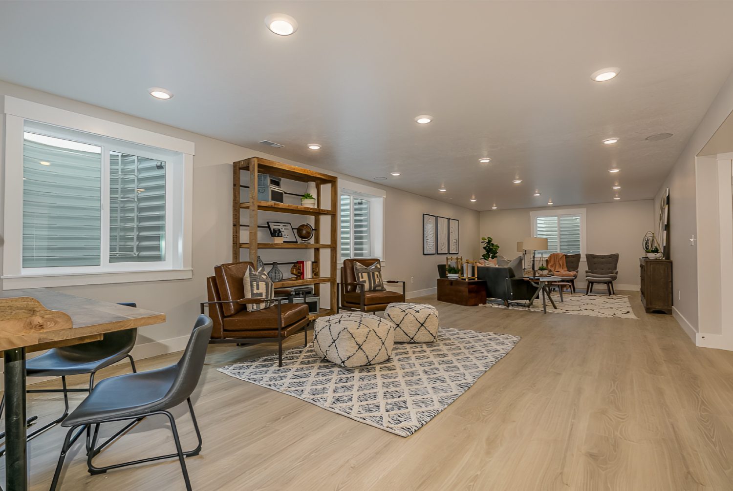 Spacious living room with recessed lighting, large windows, and wooden flooring. Features seating area with chairs, poufs, and rugs, along with bookshelves and decorative plants.