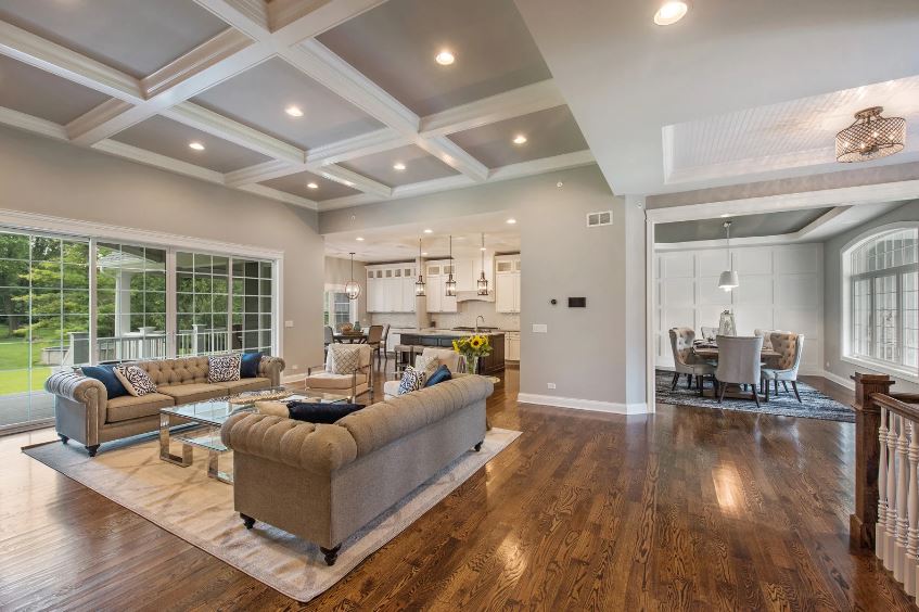 Open-concept living space with coffered ceiling, featuring a sitting area, kitchen with an island, dining area, and large glass doors leading to a green outdoor area. Hardwood floors throughout.