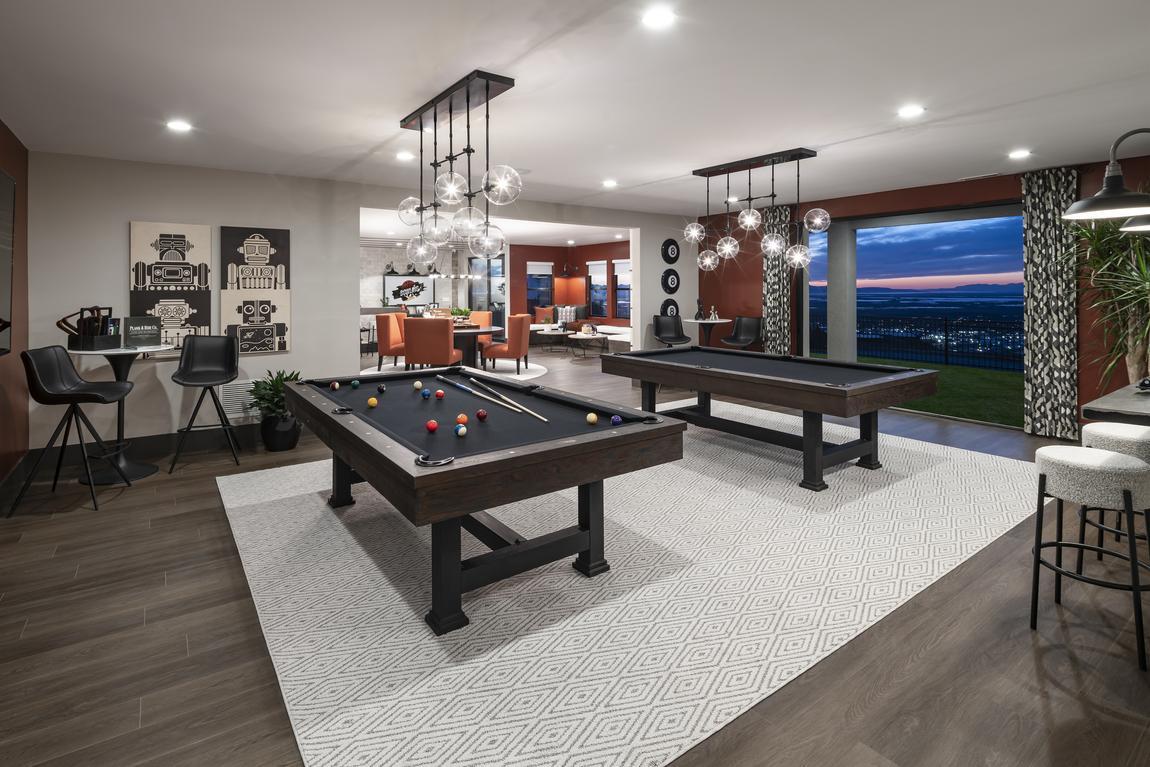 A modern game room with two pool tables, bar stools, a seating area, and decorative pendant lights. Large windows reveal an outdoor view at sunset.