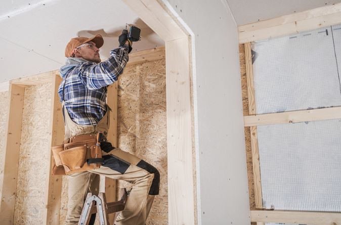 A construction worker on a ladder installs drywall in a partially framed room. He's wearing a plaid shirt, tool belt, and cap.