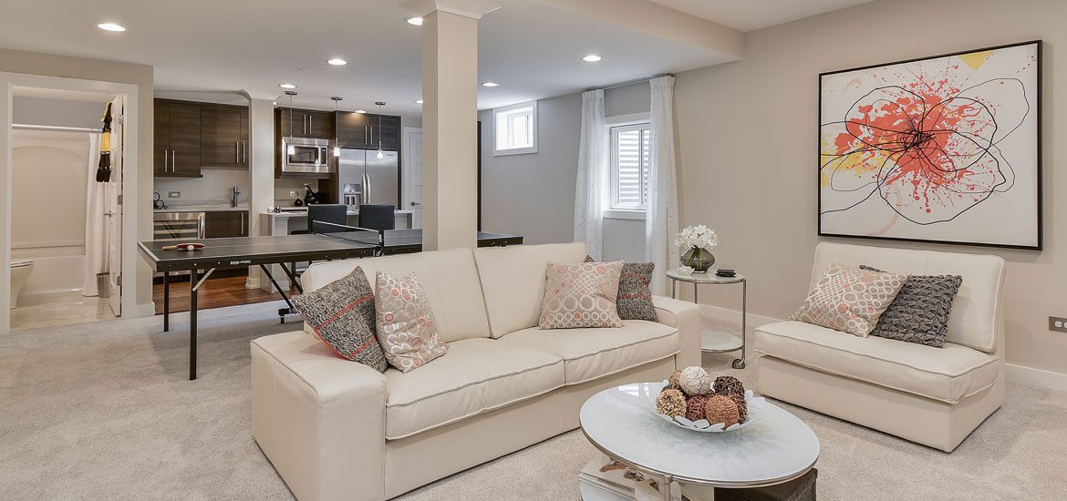 A modern living room with a beige sofa, patterned pillows, a coffee table, and abstract wall art. The open kitchen features dark wood cabinets and stainless steel appliances.