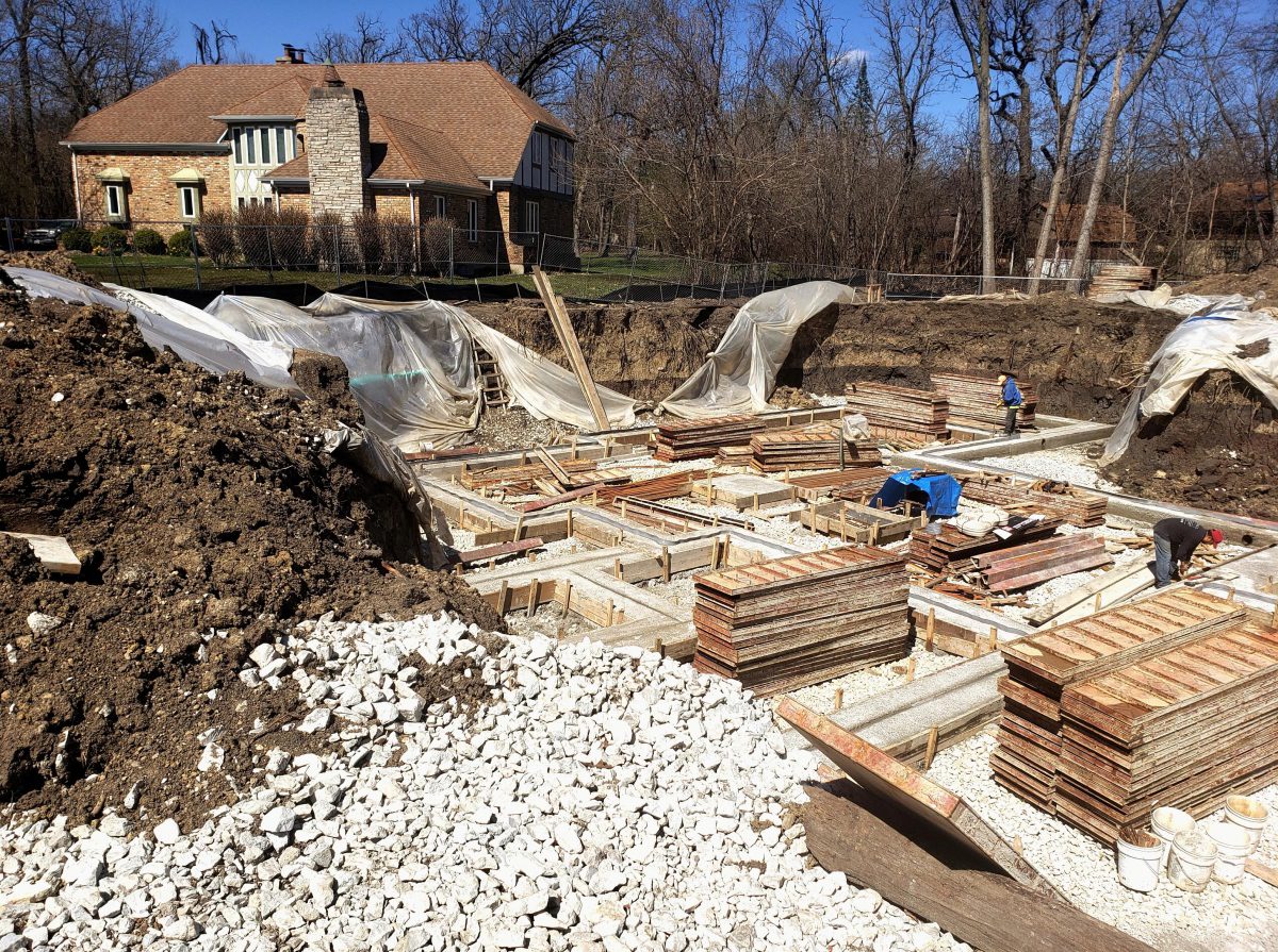 A construction site with foundation work in progress, featuring concrete forms, rebar, and gravel. A house and trees are visible in the background. Two workers are present.