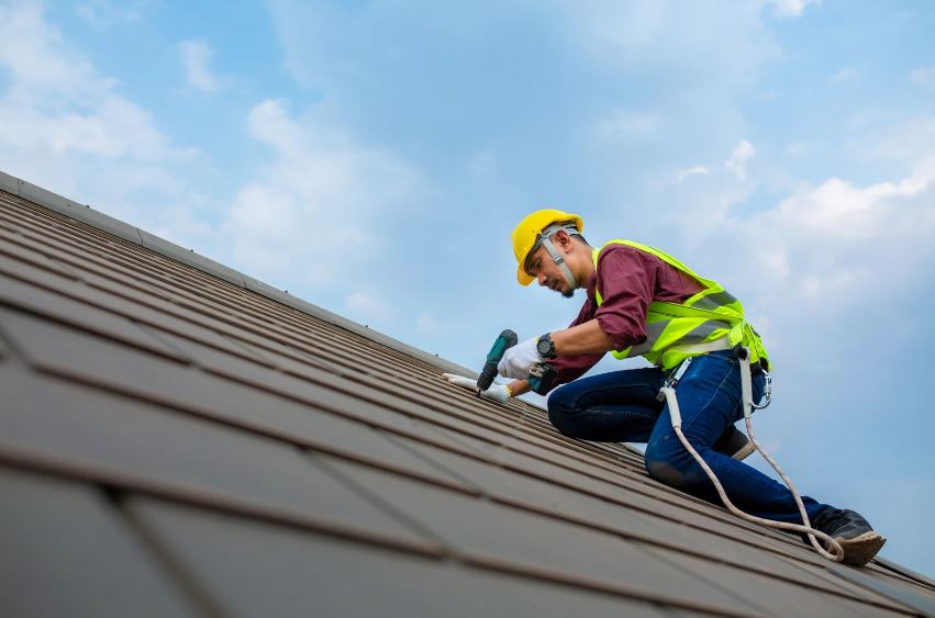 A construction worker in safety gear works on a sloped roof, using a power drill under a partly cloudy sky.