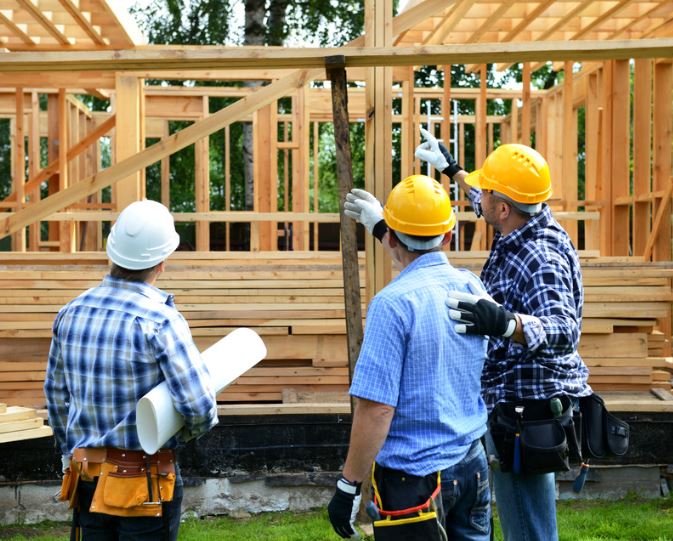 Three construction workers wearing helmets and plaid shirts discuss a wooden house frame and hold building plans.