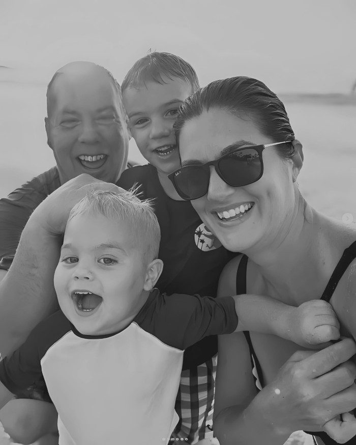 A family of four, including two young children, smiles for a selfie on a beach.